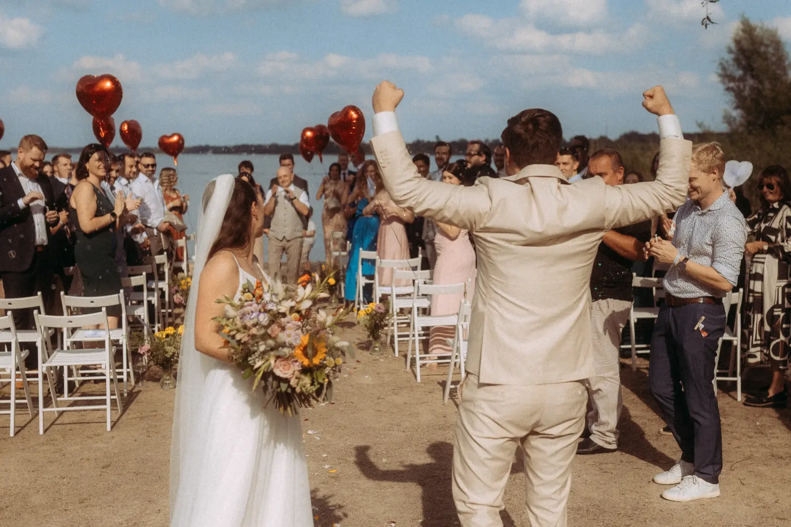 Ein glückliches Brautpaar feiert seine Trauung am See, während der Bräutigam freudig die Arme in die Luft streckt. Das Brautpaar wird von Gästen und roten Herzluftballons umgeben. Hochzeitsfotograf in Schwerin und MV.