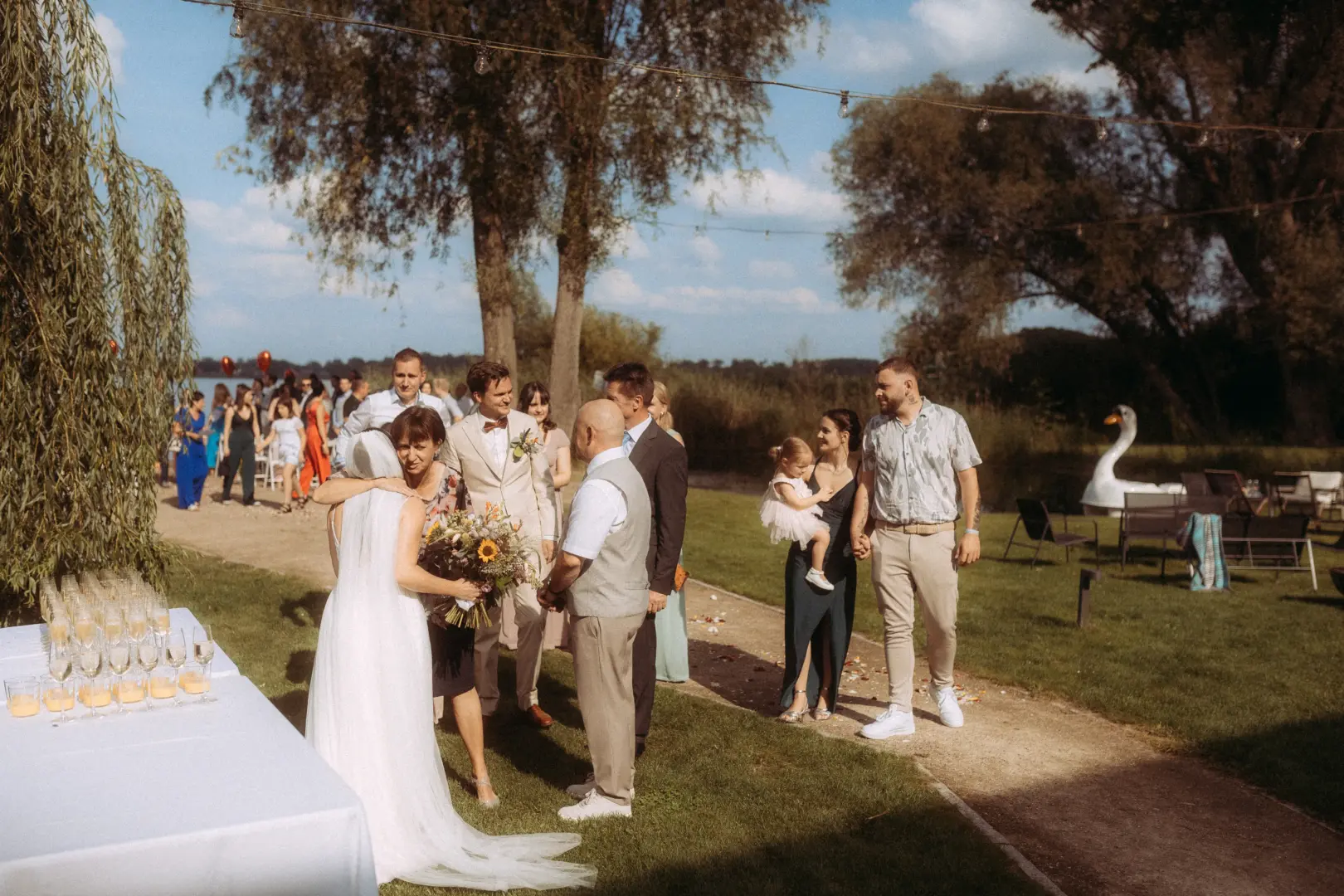 Eine Braut umarmt eine Hochzeitsgastin im Freien bei einer sommerlichen Feier am See, während Gäste im Hintergrund stehen. Das stimmungsvolle Bild fängt einen besonderen Moment ein, festgehalten von einem Hochzeitsfotograf in Schwerin und MV.