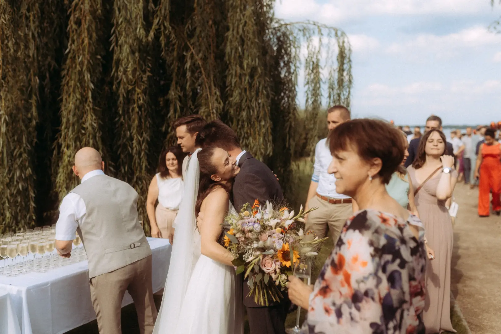 Ein Brautpaar umarmt sich herzlich bei einer Hochzeitsfeier im Freien vor einer Trauerweide, während Gäste im Hintergrund stehen. Dieses stimmungsvolle Bild zeigt die Arbeit als Hochzeitsfotograf in Schwerin und MV bei einer sommerlichen Feier am Wasser.