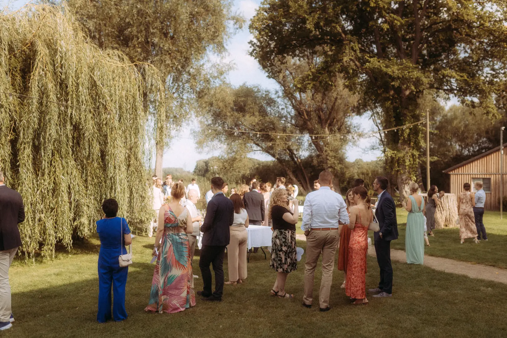 Gäste bei einer entspannten Gartenhochzeit im Grünen, festgehalten von einem Hochzeitsfotograf in Schwerin und MV. Die Hochzeitsgesellschaft in sommerlicher Kleidung unterhält sich auf einer Wiese unter großen Bäumen bei einer sommerlichen Feier im Freien.