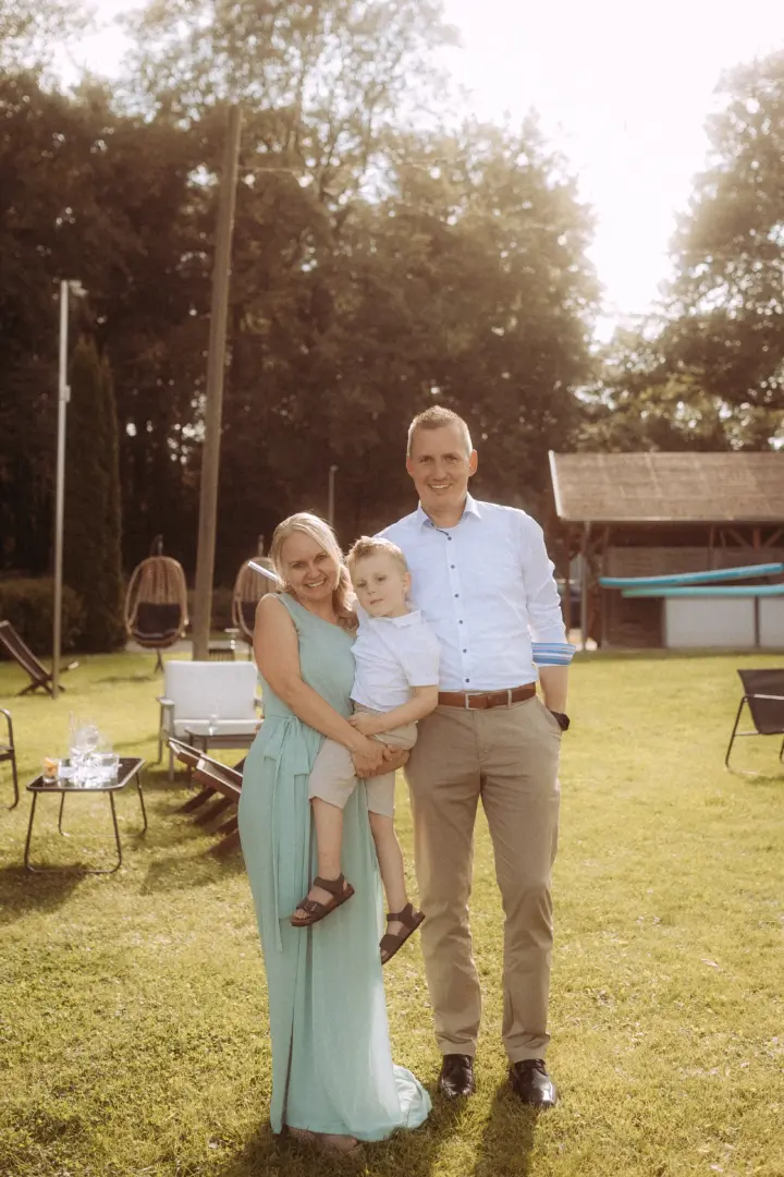 Eine glückliche Familie posiert im Garten eines Veranstaltungsortes. Die Mutter trägt ein hellgrünes Kleid, der Vater ein weißes Hemd und beigefarbene Hose, während sie ihren Sohn im Arm halten. Professioneller Familienfotograf in Schwerin für natürliche Aufnahmen im Freien.