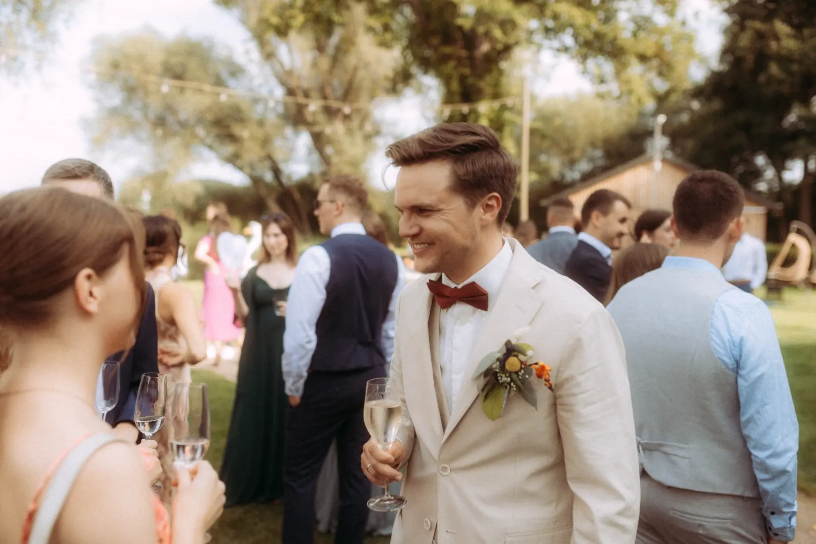 Ein lächelnder Bräutigam im beigen Anzug mit dunkelroter Fliege hält ein Glas Sekt in der Hand, während er sich auf einer sonnigen Gartenparty unterhält – festgehalten vom Hochzeitsfotograf in Schwerin und MV.