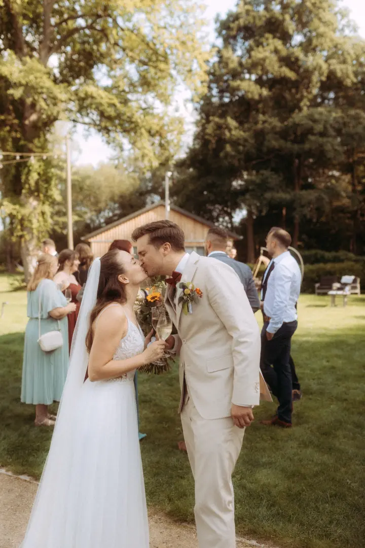 Ein Brautpaar küsst sich liebevoll bei einer sommerlichen Gartenhochzeit im Freien. Die Braut trägt ein weißes Brautkleid mit Schleier und hält einen Blumenstrauß, während der Bräutigam einen hellen Anzug trägt. Arbeit eines Hochzeitsfotograf in Schwerin und MV.