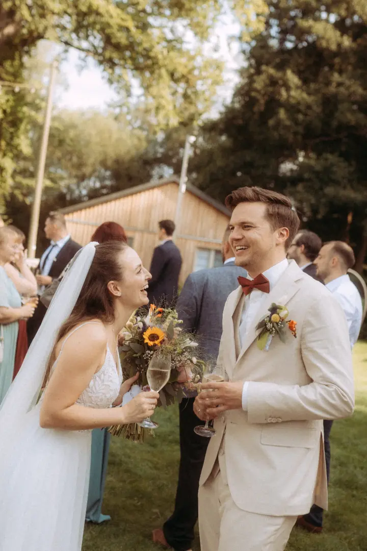 Ein glückliches Brautpaar lacht sich bei einer Gartenhochzeit an, die Braut hält einen Blumenstrauß mit Sonnenblumen und ein Weinglas. Als Lovestory Fotograf in Schwerin und MV fange ich solche authentischen Momente voller Freude fest, während Gäste im Hintergrund feiern.