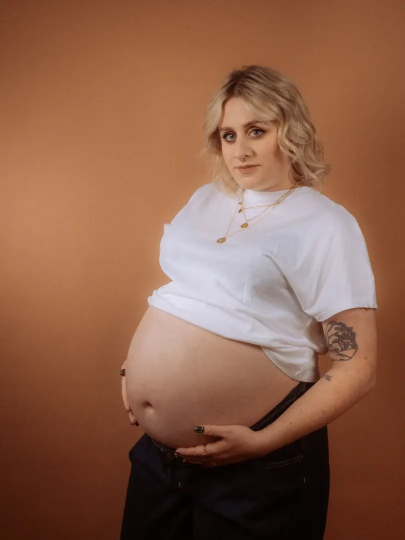 Eine schwangere Frau mit blondem Haar trägt ein weißes T-Shirt und hält liebevoll ihren Babybauch. Das Porträt wurde in einem Fotostudio in Schwerin vor einem warmen, braunen Hintergrund aufgenommen und unterstreicht die natürliche Eleganz dieser besonderen Lebensphase.