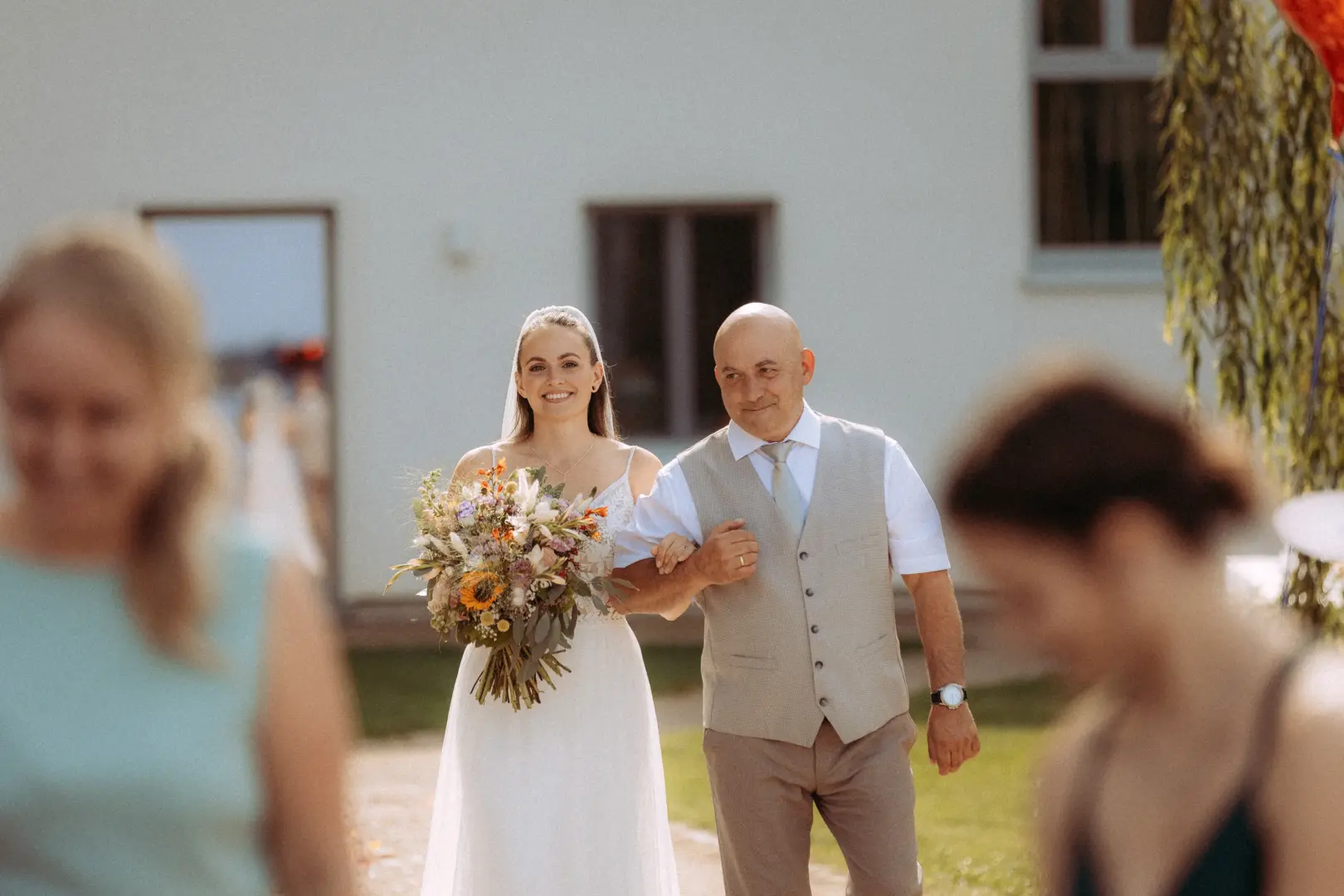 Eine strahlende Braut in einem weißen Brautkleid mit einem bunten Wildblumenstrauß wird von ihrem Vater bei der Trauung zum Altar geführt. Diese emotionale Szene wurde von einem Hochzeitsfotograf in Schwerin und MV in einem hellen, sommerlichen Ambiente festgehalten.