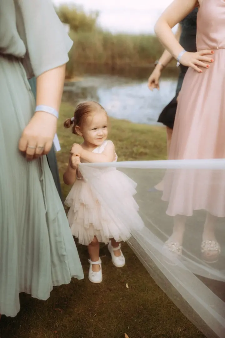Ein kleines Blumenmädchen in einem zarten, hellrosa Tüllkleid hält neugierig den Schleier der Braut, während es zwischen Gästen im Freien steht. Diese emotionale Aufnahme entstand durch einen Hochzeitsfotograf in Schwerin und MV bei einer sommerlichen Feier im Grünen.