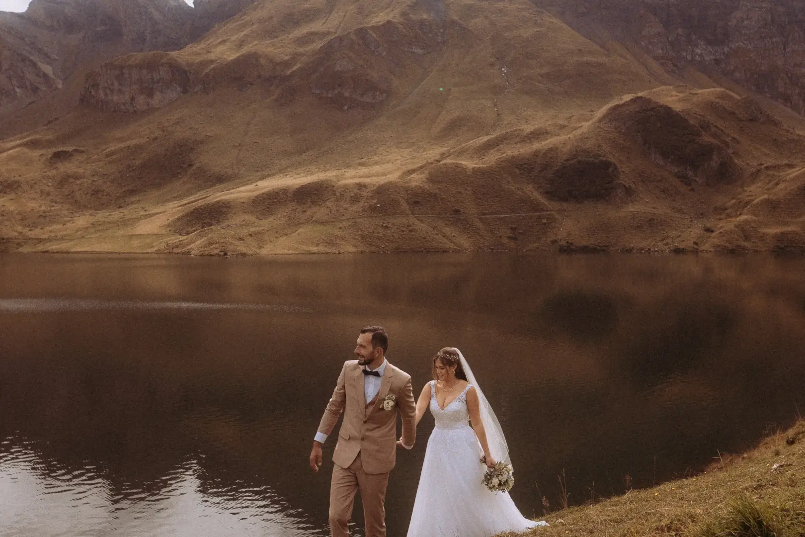 Ein Brautpaar spaziert händchenhaltend am Ufer eines ruhigen Bergsees entlang, umgeben von einer imposanten, herbstlich gefärbten Gebirgslandschaft. Dieser emotionale Moment wurde von einem kinematischen Hochzeitsfotograf in der Schweiz festgehalten.