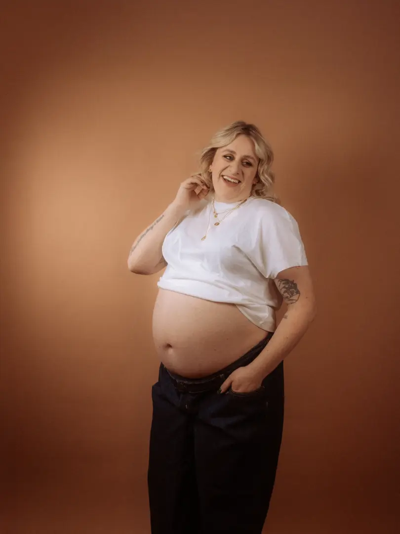 Eine strahlende schwangere Frau mit blonden Haaren lächelt im Fotostudio in Schwerin, während sie ein weißes T-Shirt trägt, das ihren Babybauch freilässt. Sie steht vor einem einfarbigen, warmen Hintergrund und hält sich eine Hand in die Tasche ihrer dunklen Jeans.