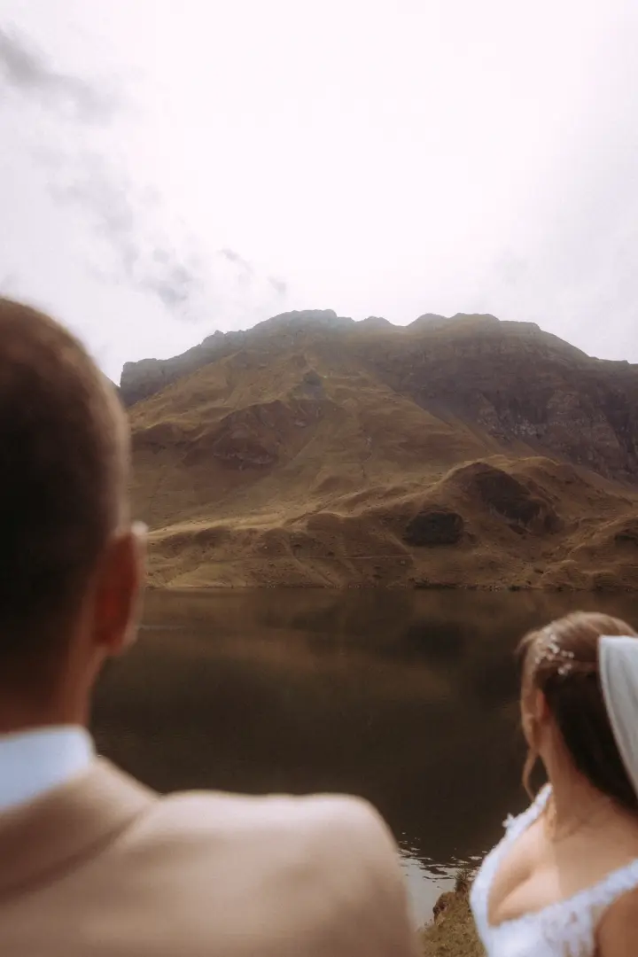 Ein Brautpaar in Hochzeitskleidung blickt gemeinsam auf einen ruhigen Bergsee vor einer majestätischen Gebirgskulisse. Diese stimmungsvolle Aufnahme zeigt den romantischen Moment als kinematischer Hochzeitsfotograf in der Schweiz.