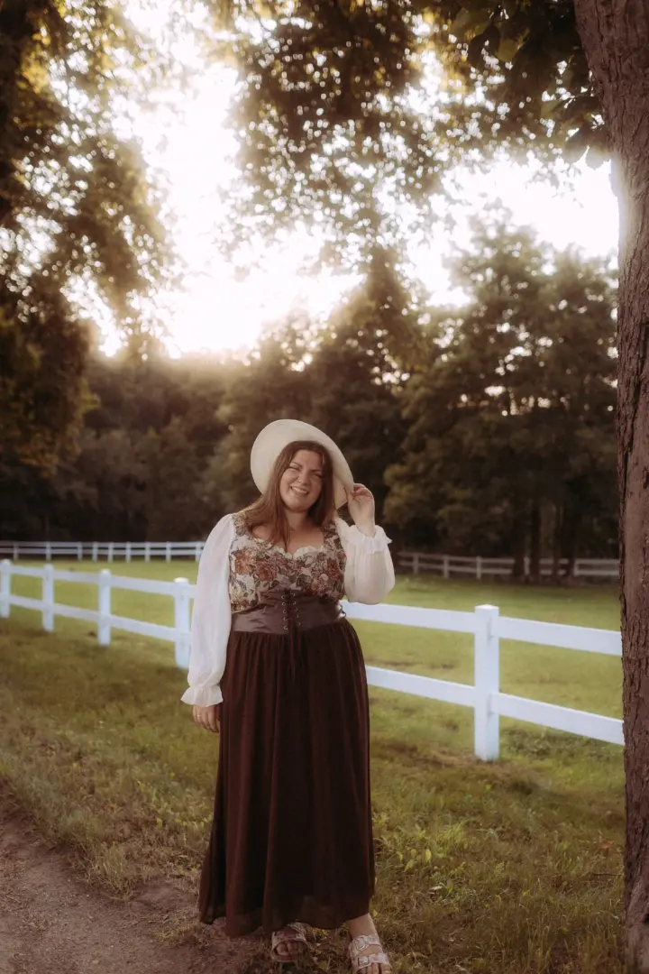 Eine lächelnde Frau in einem rustikalen, von Cottagecore inspirierten Outfit mit floralem Oberteil und braunem Rock, die einen Strohhut trägt. Sie steht bei warmem Abendlicht vor einem weißen Holzzaun auf einer Wiese. Fotograf für Pärchen in Schwerin.