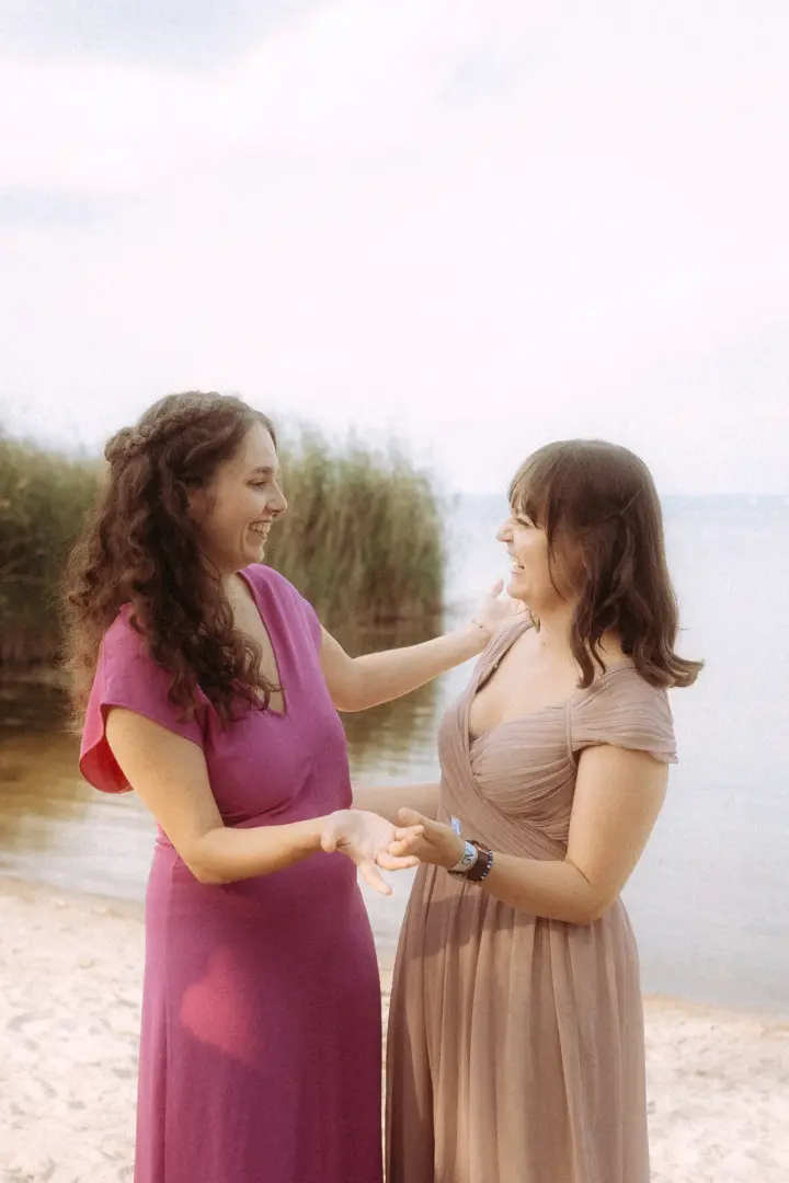 Zwei junge Frauen in eleganten Abendkleidern lachen herzlich am Strand, während sie sich an den Händen halten. Die entspannte Atmosphäre am Wasser fängt einen schönen Moment ein, fotografiert von einem Fotograf für Pärchen in Schwerin.