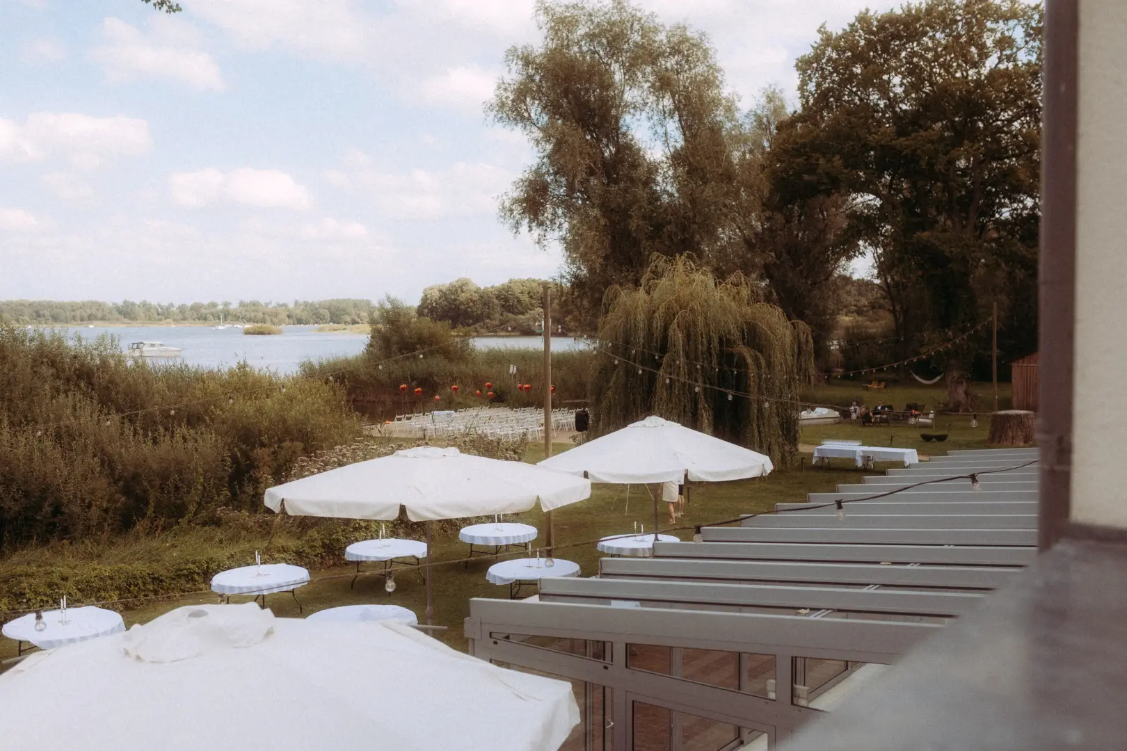 Blick von einer Terrasse auf eine Hochzeitslocation am See mit weißen Sonnenschirmen, gedeckten Tischen und Stuhlreihen für eine freie Trauung im Garten, festgehalten vom Hochzeitsfotograf Müritz und MV.