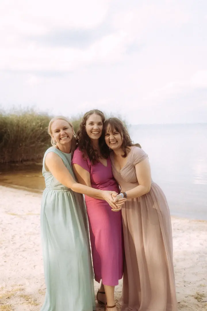 Drei glückliche Brautjungfern in pastellfarbenen Kleidern halten sich an einem sonnigen Tag am Strand an den Händen. Die Szene fängt die Freude der Hochzeit ein, fotografiert von einem Hochzeitsfotograf in Schwerin und MV.