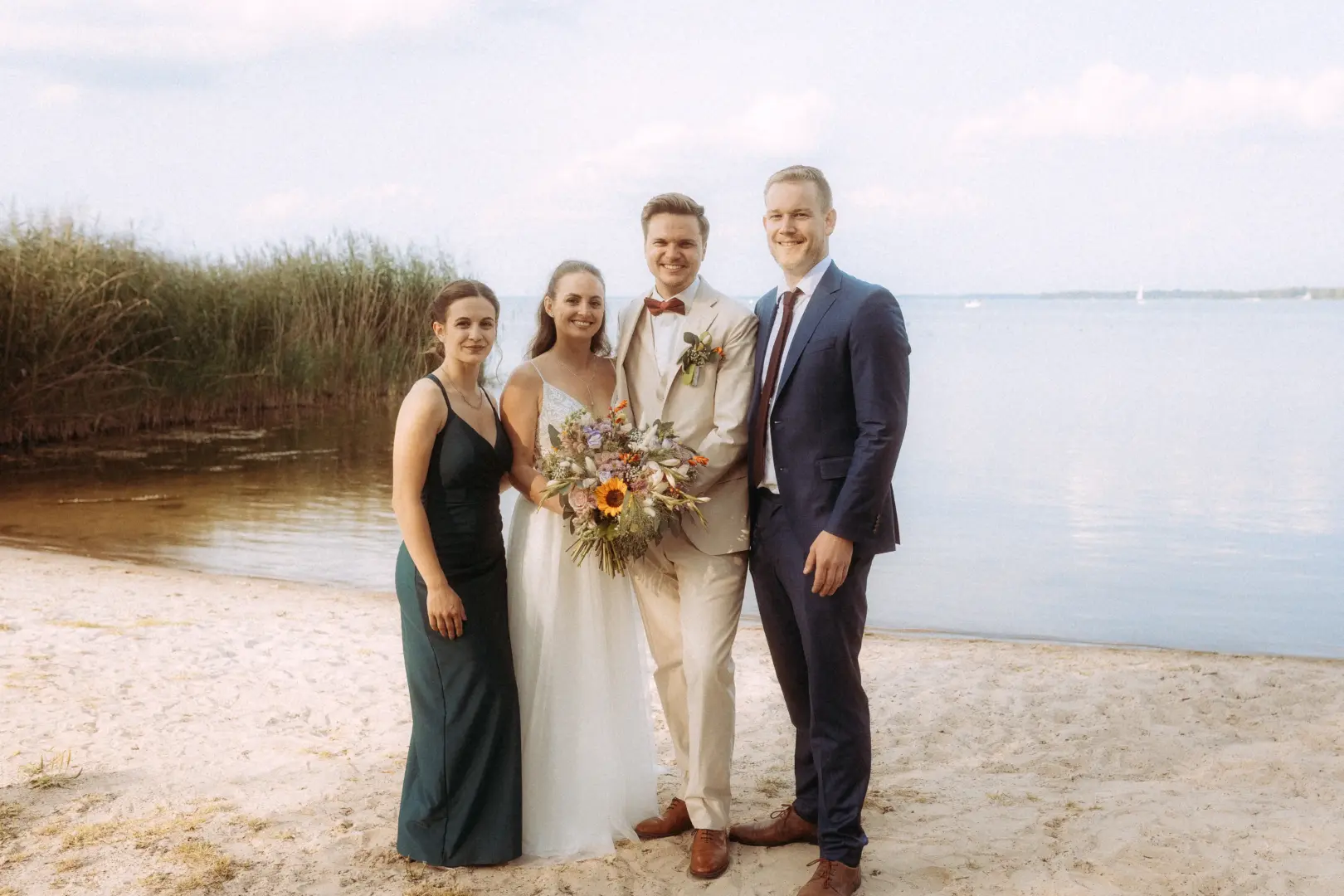 Brautpaar mit Trauzeugen posiert für ein Foto am Strand eines Sees, aufgenommen von einem Hochzeitsfotograf in Schwerin und MV. Die Braut hält einen großen Blumenstrauß, während der Bräutigam einen beigen Anzug und die Trauzeugen elegante Abendkleidung tragen.