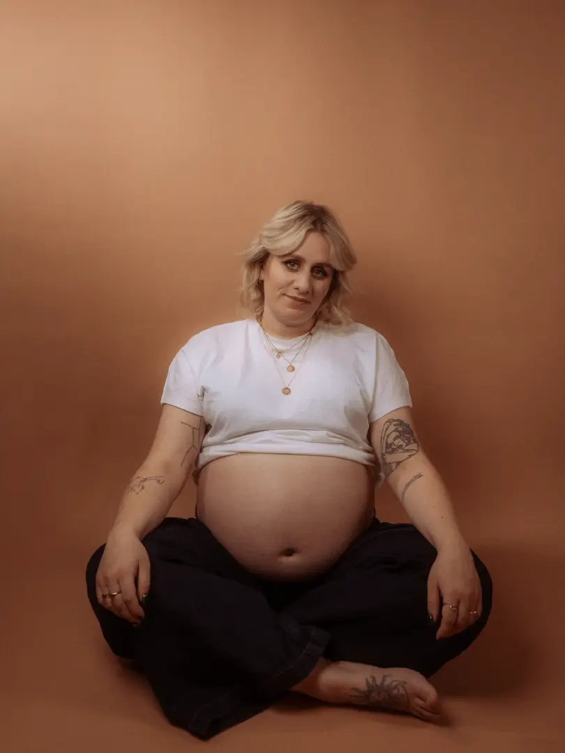 Eine schwangere Frau mit blondem Haar sitzt im Schneidersitz vor einem warmen, beigefarbenen Hintergrund. Sie trägt ein hochgeschobenes weißes T-Shirt, das ihren Babybauch betont, und dunkle Jeans. Die Aufnahme entstand in einem professionellen Fotostudio in Schwerin.