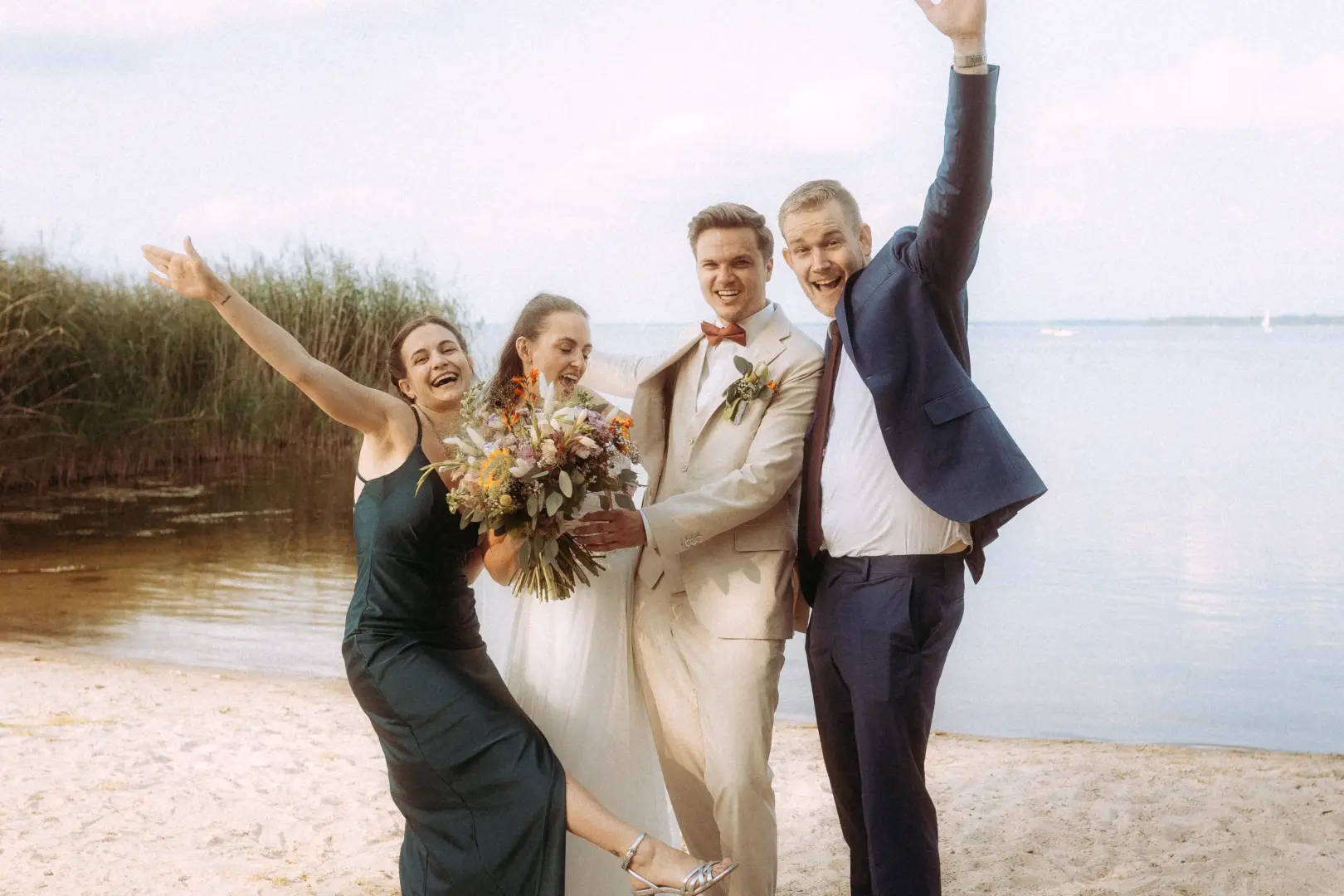 Ein glückliches Brautpaar und zwei Freunde feiern ausgelassen am Strand eines Sees. Die Braut hält einen großen Blumenstrauß, während die Gruppe fröhlich posiert. Ein authentischer Moment, festgehalten von einem professionellen Hochzeitsfotograf in Schwerin und MV.
