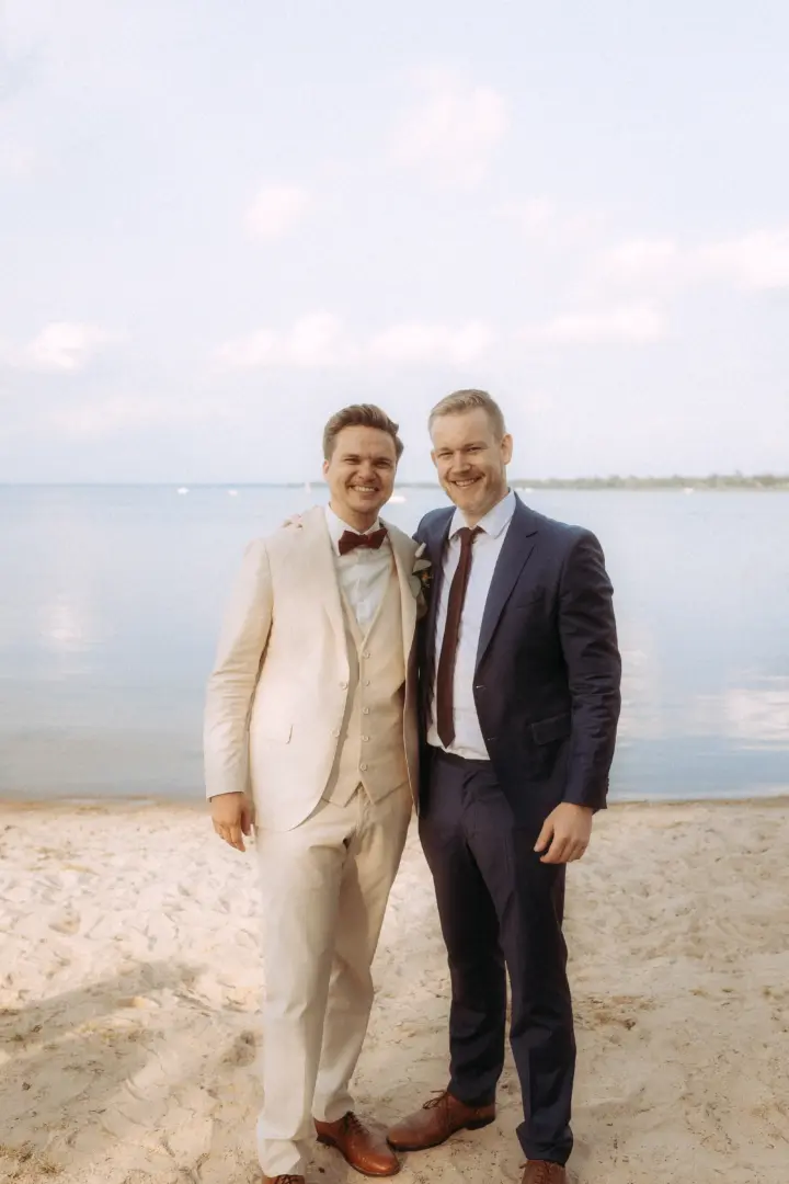 Zwei glückliche Männer in eleganten Hochzeitsanzügen stehen sich lächelnd am sandigen Ufer eines Sees gegenüber. Der Bräutigam im hellen Anzug und sein Begleiter im dunkelblauen Anzug genießen den sonnigen Tag. Professionelle Aufnahme von einem Hochzeitsfotograf in Schwerin und MV.