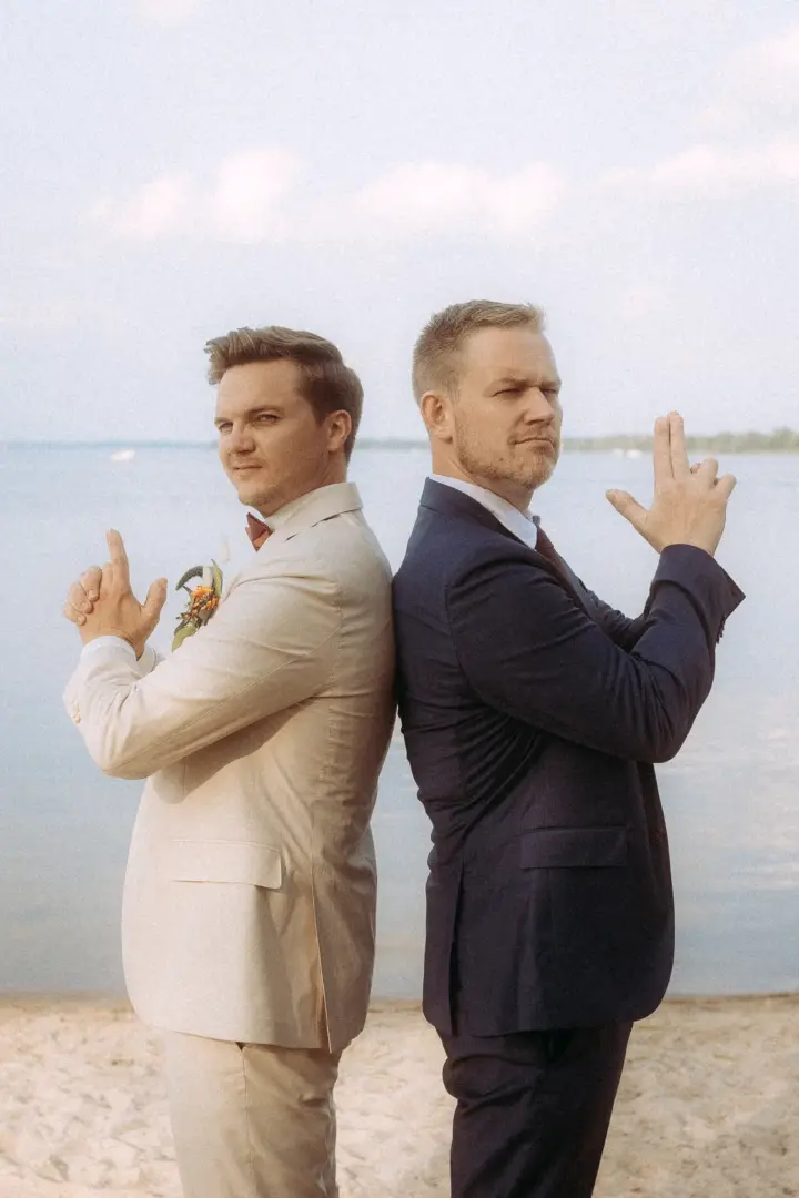 Zwei Männer in eleganten Anzügen posieren Rücken an Rücken am Strand, während sie spielerisch mit den Fingern Pistolen formen. Diese Aufnahme eines Hochzeitsfotografen in Schwerin und MV fängt die lockere, humorvolle Stimmung der beiden Bräutigame vor der ruhigen Kulisse des Sees ein.