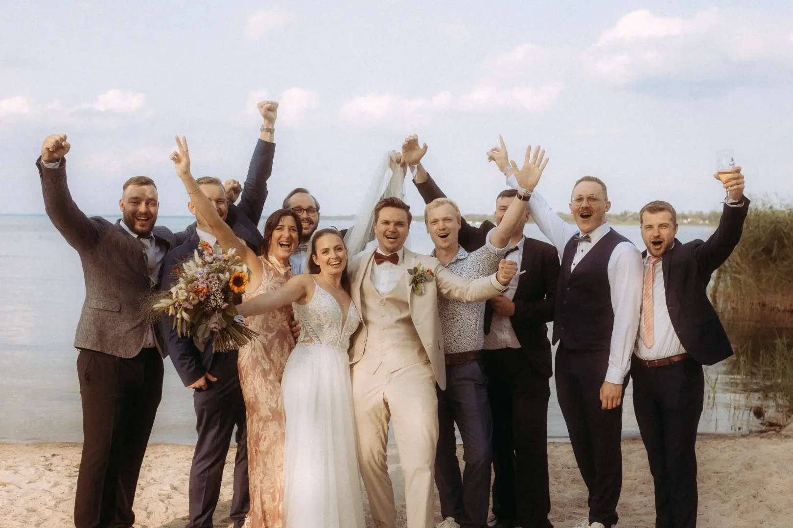 Brautpaar und Hochzeitsgesellschaft feiern ausgelassen am Strand eines Sees. Die Braut hält einen Blumenstrauß, während alle jubelnd die Arme in die Luft strecken. Professionelle Momente eingefangen von Ihrem Hochzeitsfotograf in Schwerin und MV bei einer sommerlichen Hochzeit.