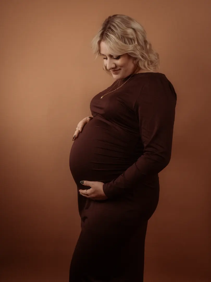 Eine schwangere Frau mit blonden Haaren trägt ein elegantes, dunkelbraunes Kleid und hält liebevoll ihren Babybauch. Die Aufnahme entstand in einem professionellen Fotostudio in Schwerin vor einem warmen, braunen Hintergrund, der eine intime und emotionale Atmosphäre schafft.