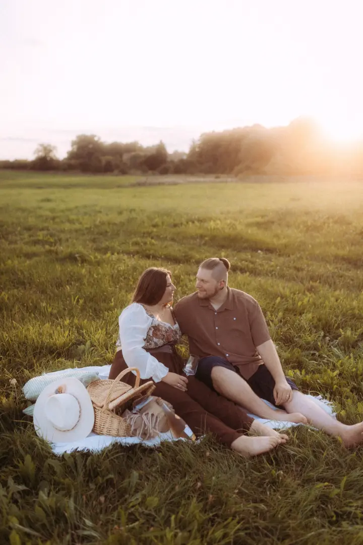 Ein verliebtes Paar sitzt bei goldenem Sonnenuntergang auf einer Decke im hohen Gras für ein romantisches Picknick. Die beiden schauen sich liebevoll an, während sie den Abend in der Natur genießen. Professionelle Pärchenfotos Schwerin, die eine intime und warme Atmosphäre einfangen.