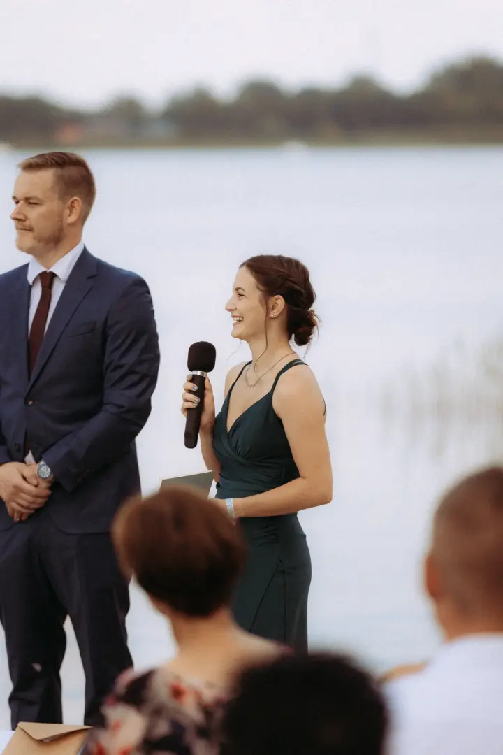 Eine junge Frau in einem eleganten, dunkelgrünen Abendkleid hält eine Rede mit einem Mikrofon während einer Hochzeitsfeier am Wasser. Neben ihr steht ein Mann im dunkelblauen Anzug. Ein stimmungsvoller Moment, eingefangen von Ihrem Hochzeitsfotograf in Schwerin und MV.