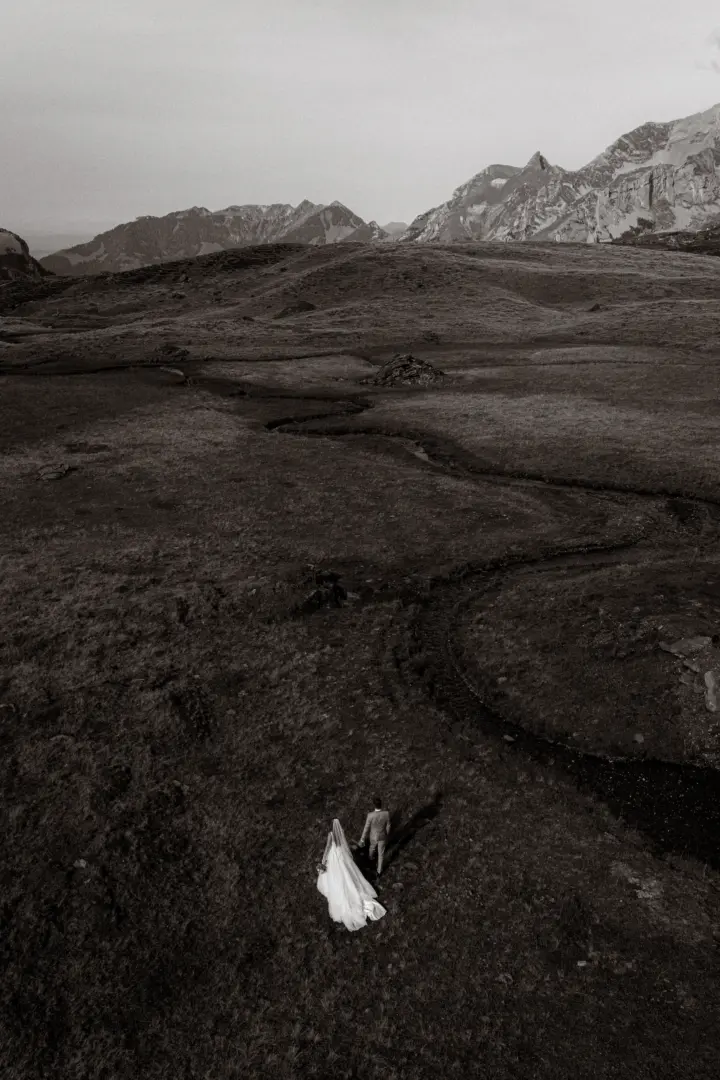 Brautpaar in einem weißen Hochzeitskleid und Anzug spaziert Hand in Hand durch eine weite, dramatische Alpenlandschaft mit einem kleinen Bachlauf und majestätischen Bergen im Hintergrund. Kinematischer Hochzeitsfotograf in der Schweiz.