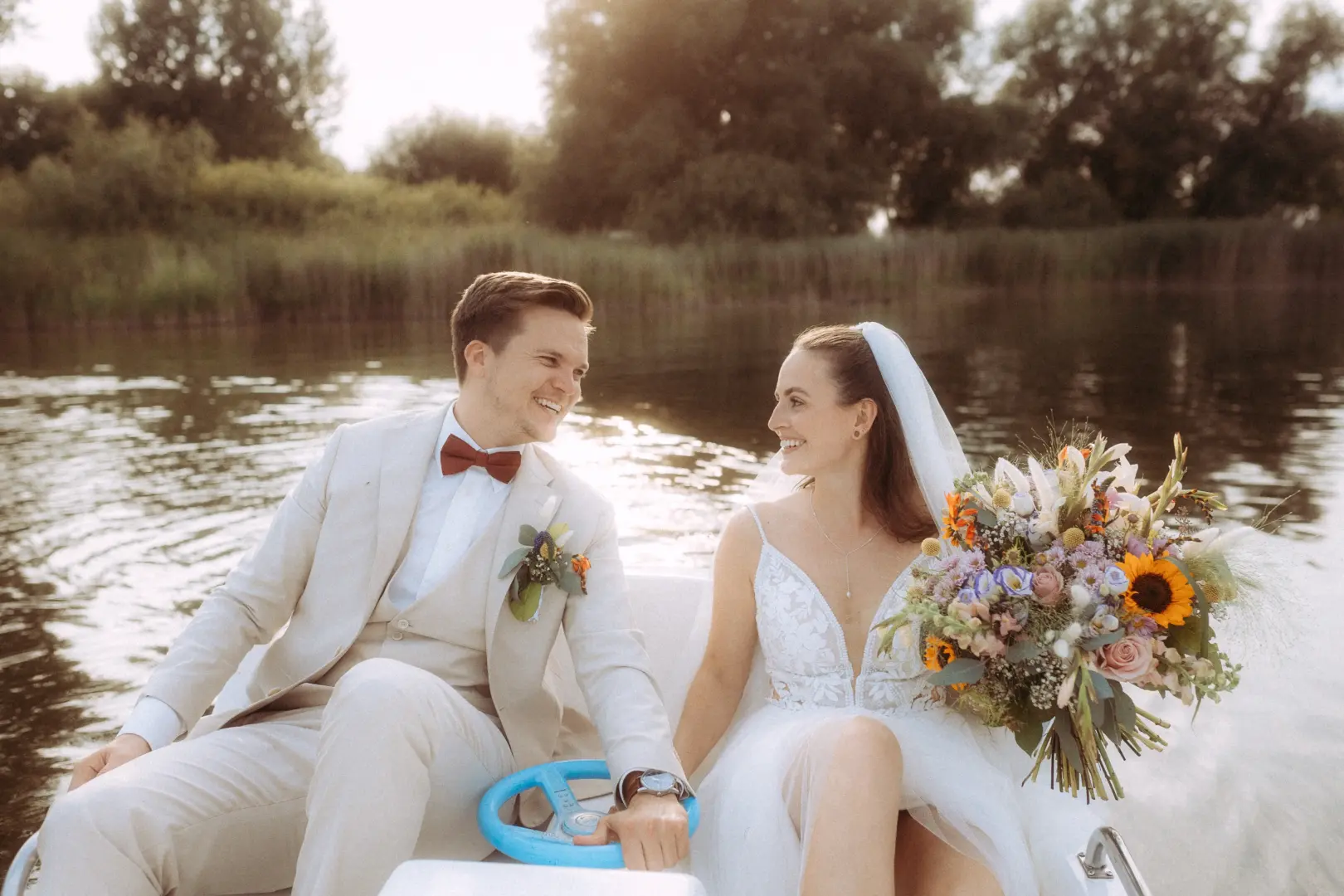 Ein glückliches Brautpaar bei einer romantischen Bootsfahrt auf einem See, festgehalten von einem Hochzeitsfotograf in Schwerin und MV. Die Braut hält einen bunten Blumenstrauß, während sie sich im hellen Hochzeitsanzug und weißen Kleid anlächeln, umgeben von idyllischer Natur.