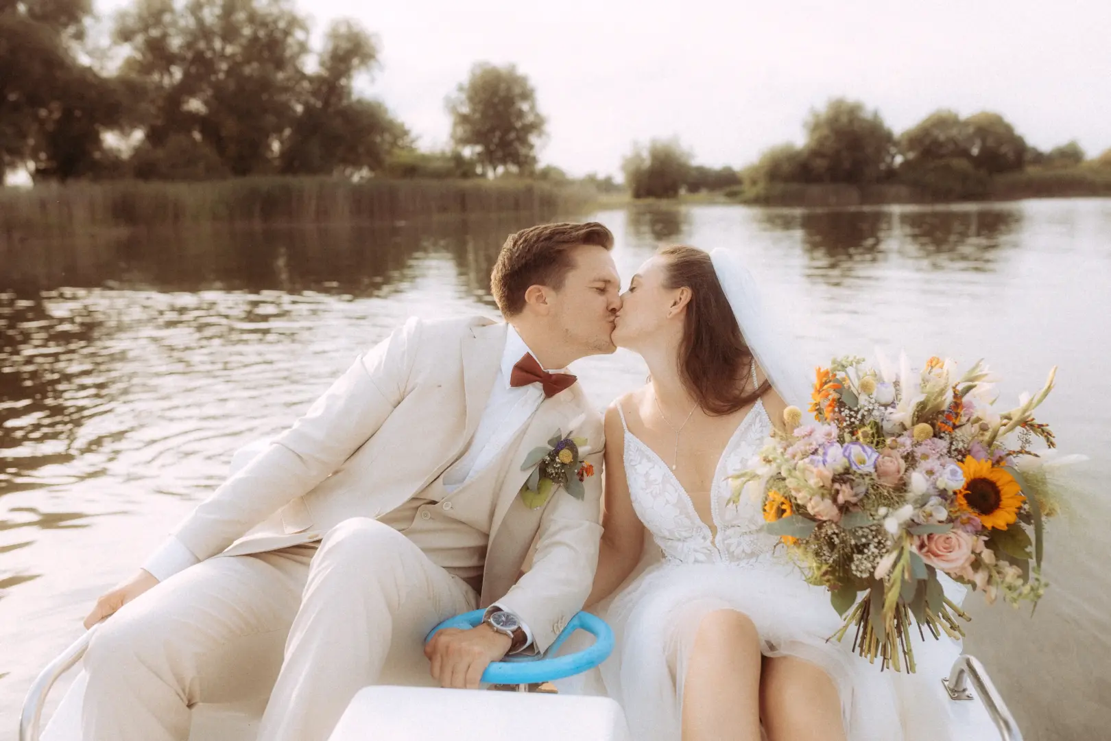 Ein glückliches Brautpaar küsst sich romantisch auf einem Tretboot auf einem ruhigen See. Die Braut hält einen großen, bunten Blumenstrauß mit Sonnenblumen, während der Bräutigam im hellen Anzug das Steuer hält. Ein stimmungsvolles Bild vom Hochzeitsfotograf in Schwerin und MV.