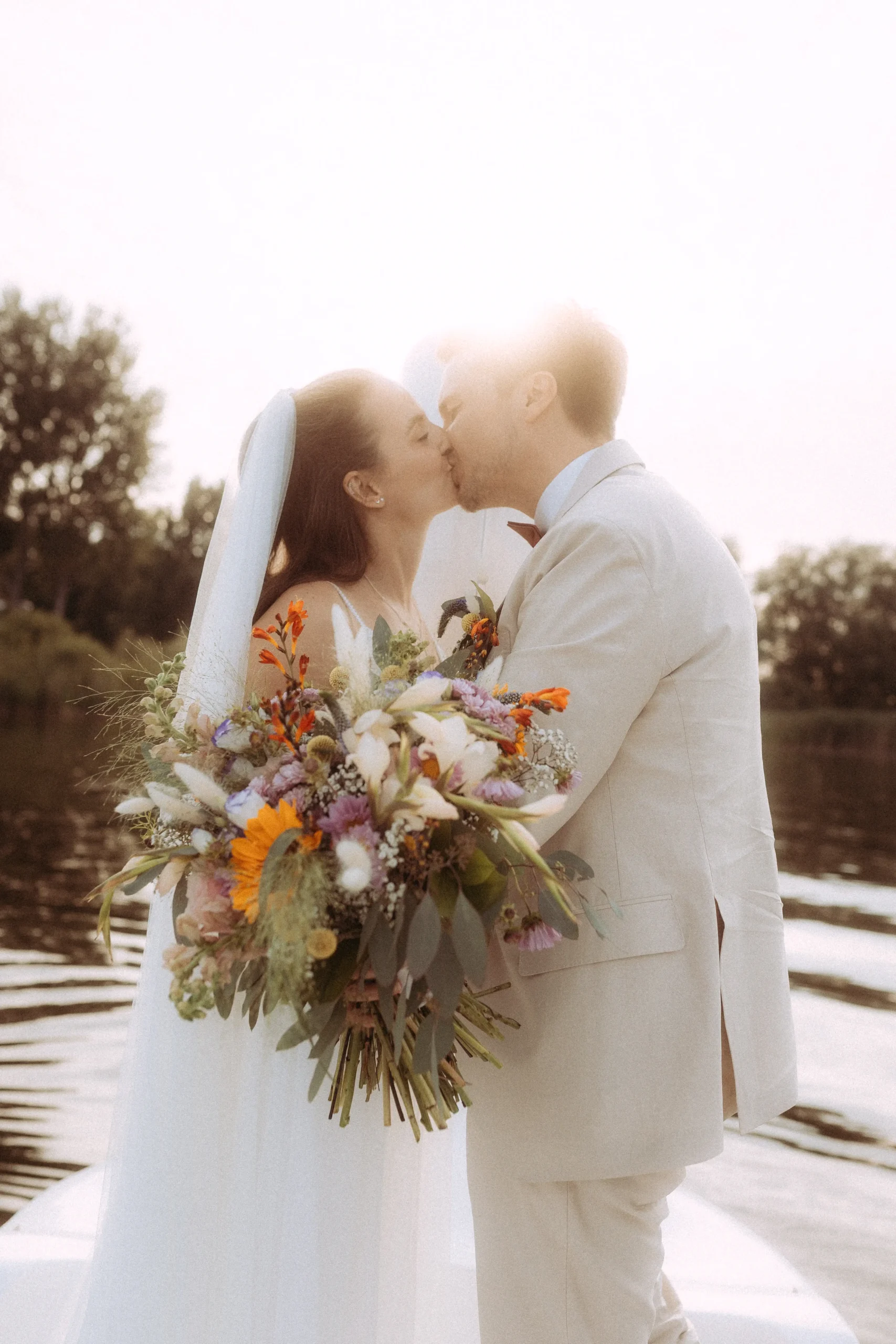 Ein frisch vermähltes Brautpaar küsst sich zärtlich am Ufer eines Sees bei Sonnenuntergang. Die Braut hält einen großen, farbenfrohen Wildblumenstrauß. Romantische Hochzeitsfotografie von einem Hochzeitsfotograf in Schwerin und MV für unvergessliche Momente.