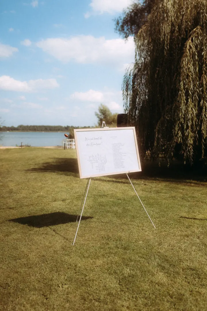 Ein auf einer Staffelei stehendes Hochzeitsrätsel mit der Aufschrift „Wie gut kennst du das Brautpaar?“ auf einer sonnigen Wiese am Seeufer. Im Hintergrund sorgt eine Trauerweide für eine idyllische Kulisse, eingefangen von einem Hochzeitsfotograf in Schwerin und MV.