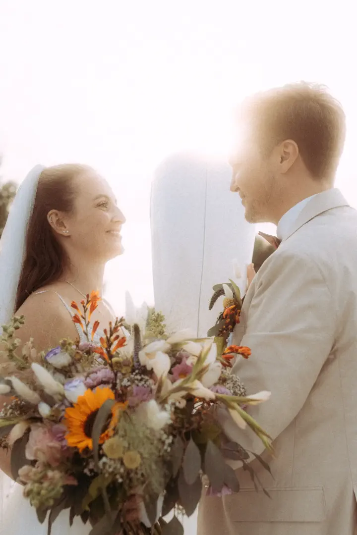 Ein glückliches Brautpaar blickt sich bei strahlendem Sonnenschein liebevoll an, während die Braut einen bunten Blumenstrauß mit Sonnenblumen hält. Diese romantische Aufnahme wurde von einem Hochzeitsfotograf in Schwerin und MV festgehalten.