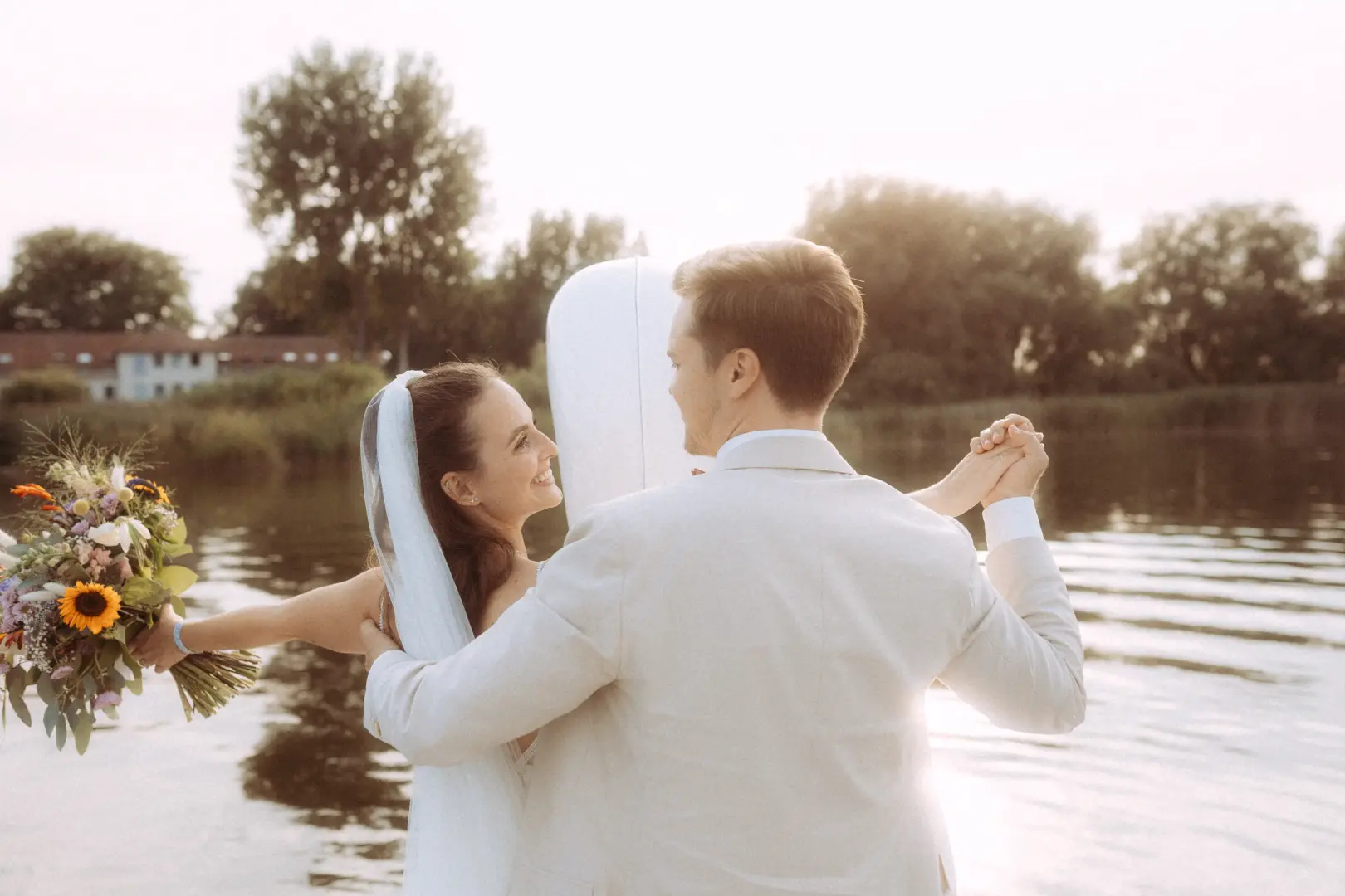 Ein glückliches Brautpaar tanzt bei Sonnenuntergang am See, während die Braut einen bunten Blumenstrauß mit Sonnenblumen hält. Diese romantische Aufnahme wurde von einem Hochzeitsfotograf in Schwerin und MV festgehalten.