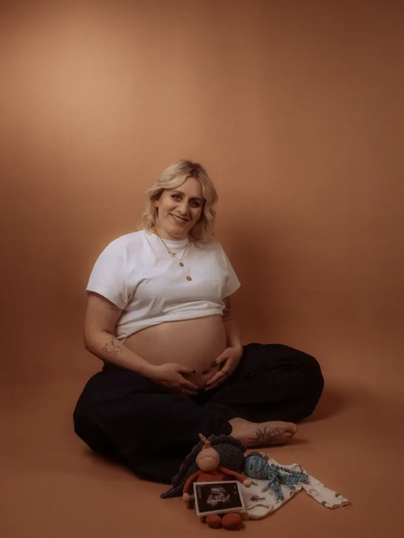 Eine lächelnde, strahlende schwangere Frau sitzt auf dem Boden und berührt ihren Bauch. Sie trägt ein weißes T-Shirt und sitzt vor einem warmen, braunen Hintergrund. Vor ihr liegen ein Baby-Ultraschallbild, ein Plüschtier und Babykleidung.