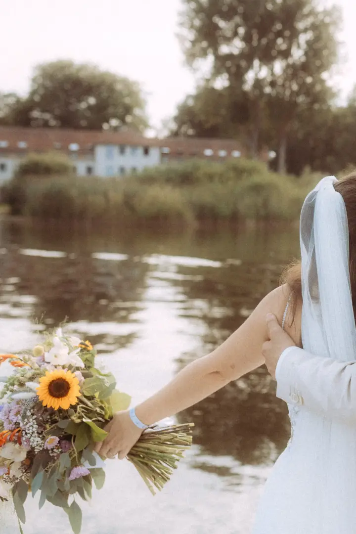 Eine Braut im weißen Kleid mit einem bunten Brautstrauß aus Sonnenblumen hält die Hand ihres Partners am Ufer eines Sees. Die romantische Stimmung fängt einen intimen Moment ein, perfekt festgehalten durch einen Hochzeitsfotograf in Schwerin und MV.