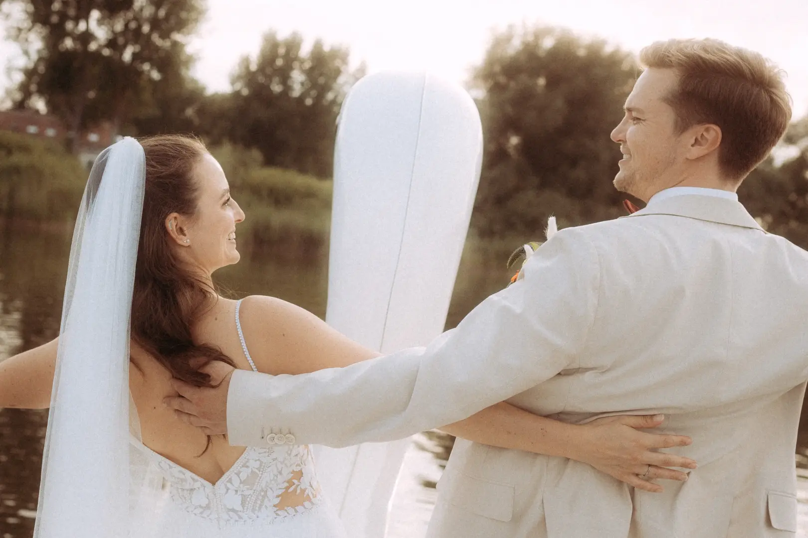 Ein glückliches Brautpaar blickt sich bei einem romantischen Fotoshooting am Wasser liebevoll in die Augen. Die Braut trägt ein elegantes Spitzenkleid mit Schleier, der Bräutigam einen hellen Anzug. Eine stimmungsvolle Aufnahme vom Hochzeitsfotograf in Schwerin und MV.