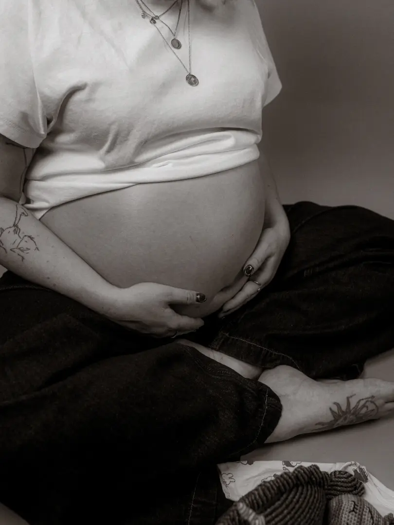 Eine schwangere Frau im Fotostudio in Schwerin hält liebevoll ihren Babybauch, während sie entspannt im Schneidersitz auf dem Boden sitzt. Sie trägt ein schlichtes weißes T-Shirt und eine dunkle Hose, ihre Arme sind mit Tattoos verziert, und neben ihr liegen kleine Babysachen.