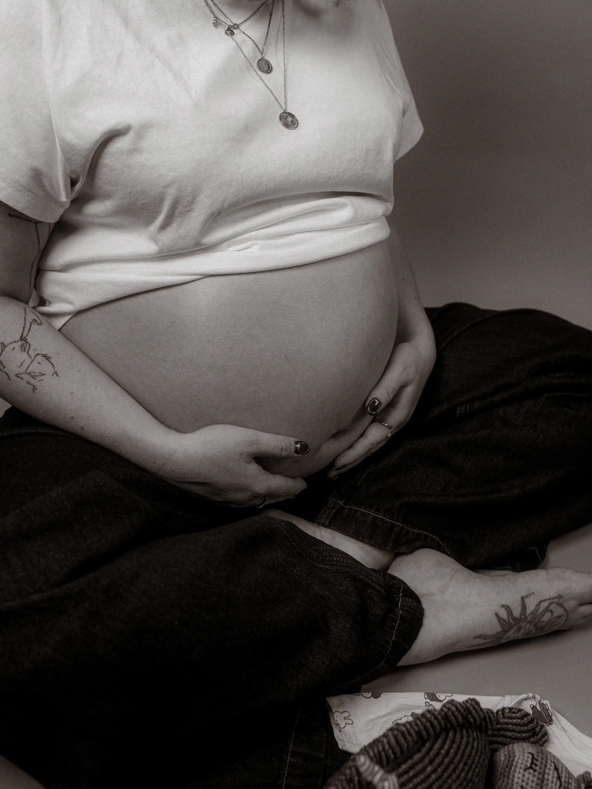 Eine schwangere Frau im Fotostudio in Schwerin hält liebevoll ihren Babybauch, während sie entspannt im Schneidersitz auf dem Boden sitzt. Sie trägt ein schlichtes weißes T-Shirt und eine dunkle Hose, ihre Arme sind mit Tattoos verziert, und neben ihr liegen kleine Babysachen. Eine schwangere Frau im Fotostudio in Schwerin hält liebevoll ihren Babybauch, während sie entspannt im Schneidersitz auf dem Boden sitzt. Sie trägt ein schlichtes weißes T-Shirt und eine dunkle Hose, ihre Arme sind mit Tattoos verziert, und neben ihr liegen kleine Babysachen.