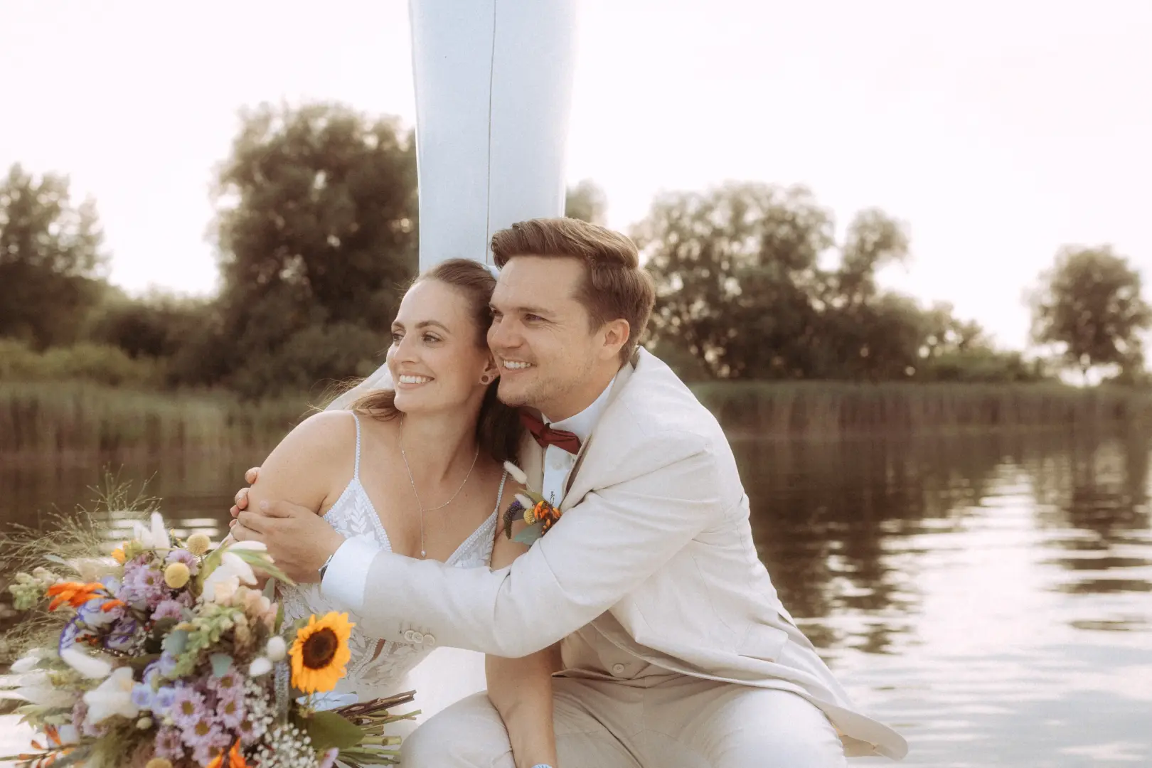 Ein glückliches Brautpaar bei einem romantischen Fotoshooting am Wasser: Die Braut im weißen Spitzenkleid hält einen bunten Blumenstrauß, während der Bräutigam im hellen Anzug sie liebevoll umarmt. Professionelle Lovestory Fotografen in Schwerin und MV halten diesen intimen Moment fest.