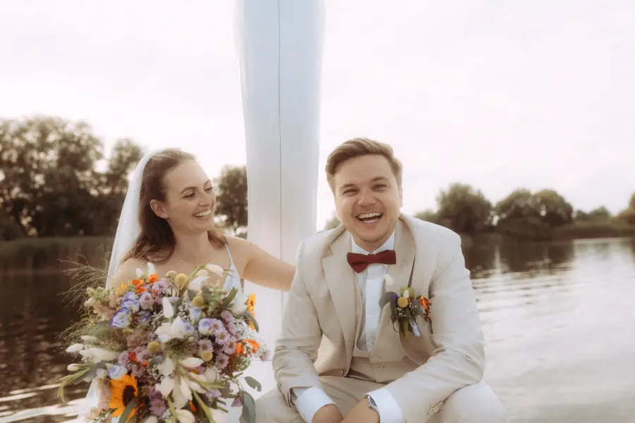 Ein glückliches Brautpaar lacht gemeinsam auf einem Boot vor einer idyllischen Seenlandschaft. Die Braut hält einen bunten Blumenstrauß, während der Bräutigam einen hellen Anzug trägt. Authentische Hochzeitsfotograf in Schwerin und MV Aufnahme eines verliebten Paares an einem sonnigen Tag.