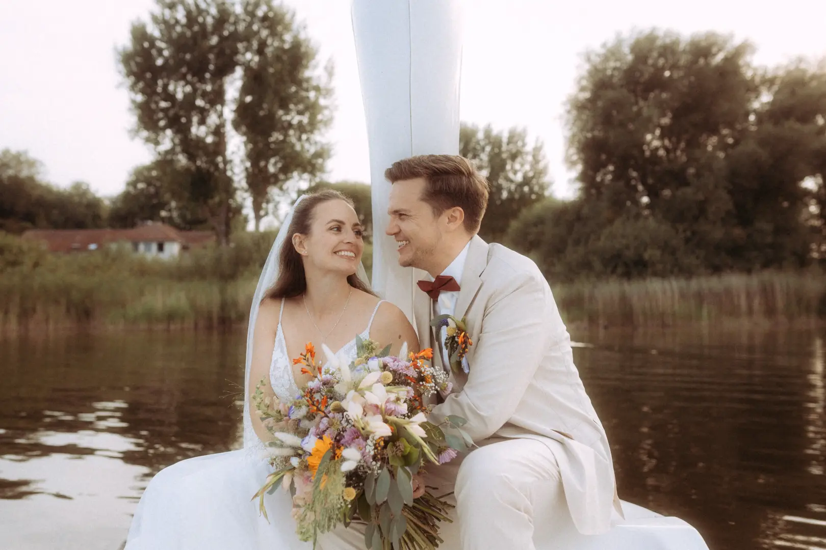 Ein glückliches Brautpaar sitzt eng umschlungen auf einem Boot vor einer malerischen Seenlandschaft. Die Braut hält einen bunten Wildblumenstrauß, während der Bräutigam im beigen Anzug sie liebevoll ansieht. Ein authentischer Moment, festgehalten vom Hochzeitsfotograf in Schwerin und MV.