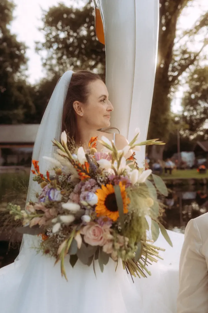 Eine Braut mit Schleier blickt verträumt in die Ferne und hält einen großen, farbenfrohen Brautstrauß mit Sonnenblumen. Die Aufnahme besticht durch eine romantische Stimmung am Wasser und wurde von einem Hochzeitsfotograf in Schwerin und MV festgehalten.