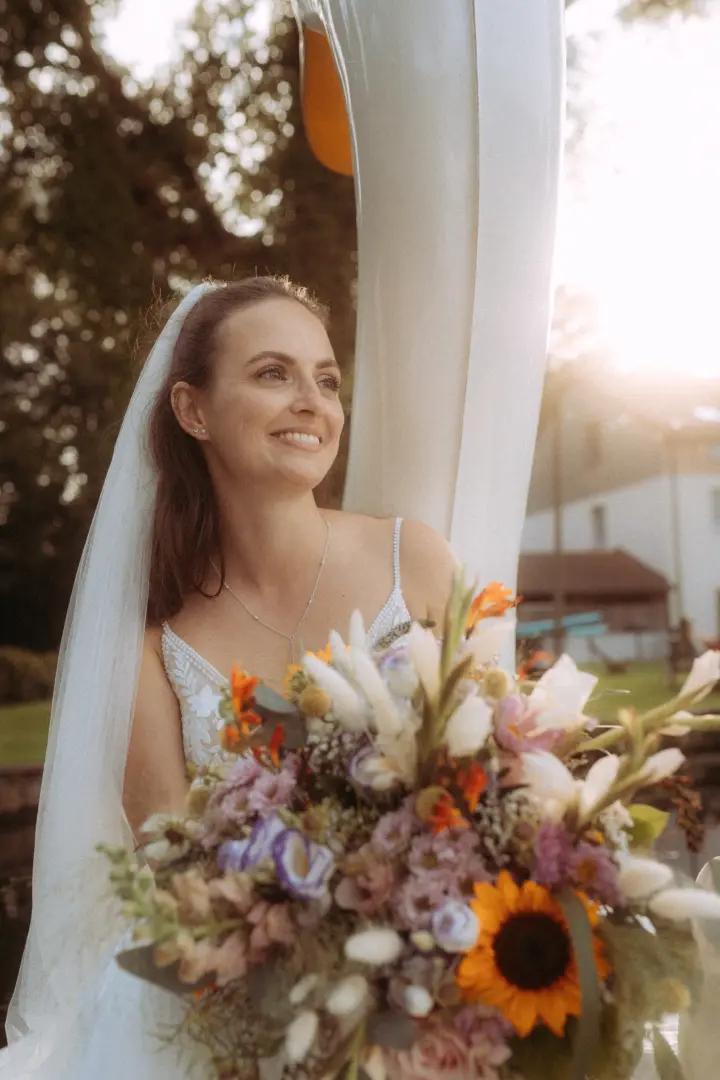 Eine strahlende Braut in einem weißen Hochzeitskleid mit Schleier hält einen farbenfrohen Blumenstrauß mit Sonnenblumen in den Händen, während sie im warmen Licht der Abendsonne lächelt – festgehalten von deinem Hochzeitsfotograf in Schwerin und MV.