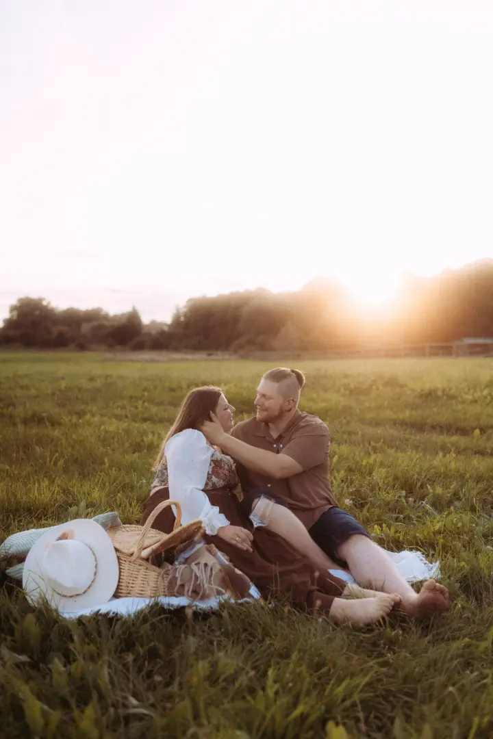 Ein verliebtes Paar bei einem romantischen Picknick auf einer Wiese im goldenen Licht der untergehenden Sonne. Der Mann hält zärtlich das Gesicht der Frau, während sie sich ansehen. Stimmungsvolle Pärchenfotos Schwerin in natürlicher Umgebung mit Picknickkorb und Strohhut.