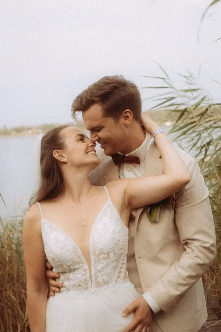 Ein glückliches Brautpaar in romantischer Pose am Seeufer, aufgenommen von einem Hochzeitsfotograf in Schwerin und MV. Die Braut trägt ein weißes Spitzenkleid, der Bräutigam einen hellen Anzug mit Fliege, während sie sich liebevoll ansehen und die Naturkulisse genießen.