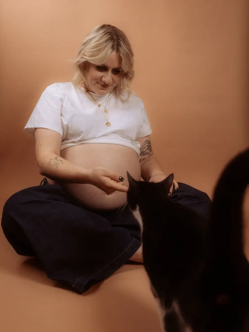 Eine schwangere Frau mit blondem Haar sitzt im Fotostudio in Schwerin auf dem Boden, trägt ein weißes T-Shirt und eine dunkle Jeans, während sie liebevoll ihre Hand nach einer schwarz-weißen Katze ausstreckt.