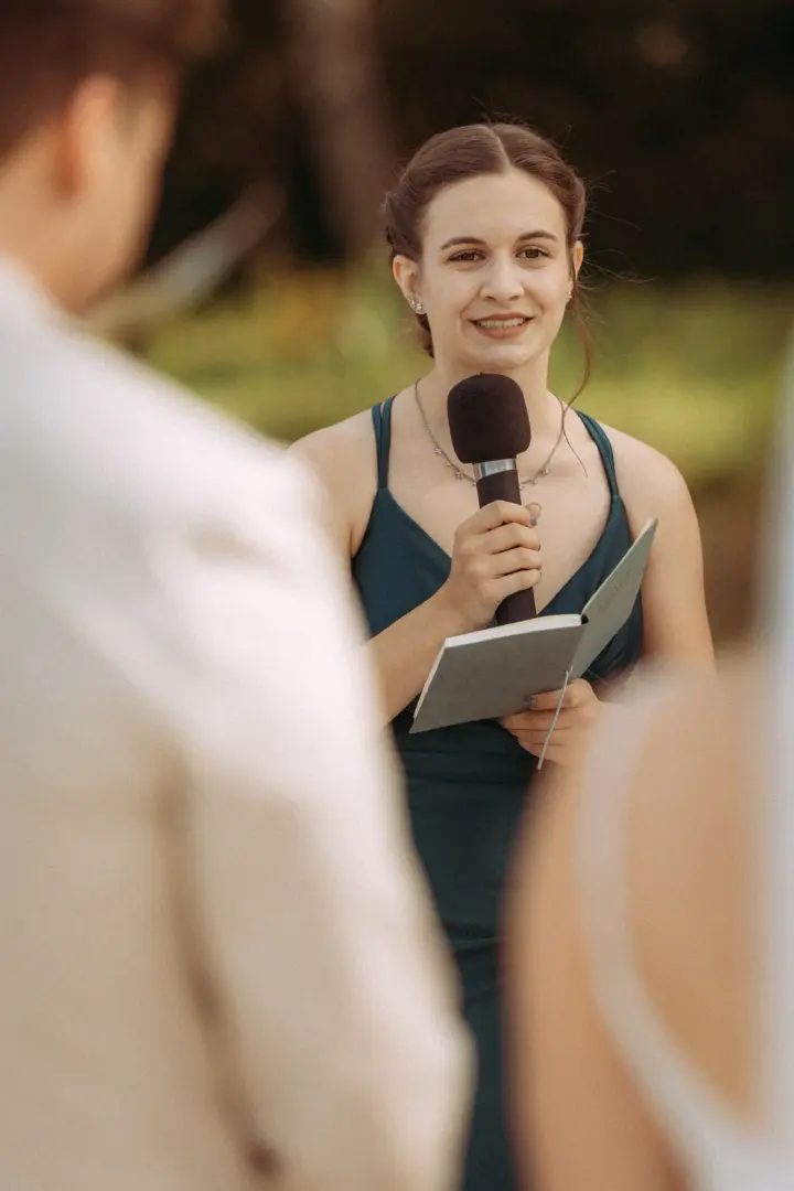 Eine junge Frau in einem eleganten dunkelgrünen Kleid hält während einer emotionalen Zeremonie eine Rede in ein Mikrofon und liest aus einem kleinen Notizbuch vor, festgehalten von einem Hochzeitsfotograf in Schwerin und MV.
