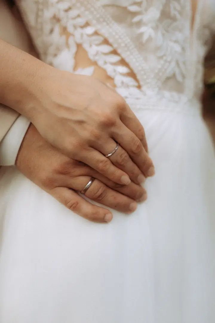 Nahaufnahme der Hände eines Brautpaares, die sich zärtlich auf dem weißen Hochzeitskleid mit floralen Spitzenapplikationen halten, wobei die Eheringe deutlich sichtbar sind. Romantische Aufnahme für eine Lovestory vom Fotograf für Pärchen in Schwerin.