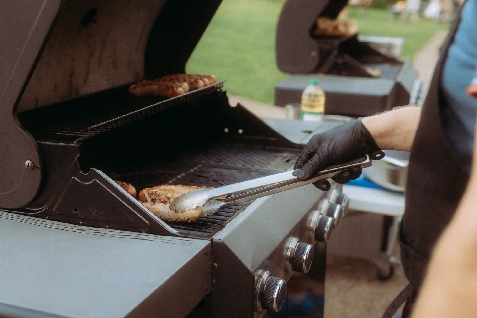 Eine Person mit schwarzem Handschuh wendet mit einer Grillzange Bratwürste auf einem Gasgrill. Im Hintergrund ist ein weiterer Grill und eine grüne Wiese zu sehen, während das Catering für ein Event vorbereitet wird.
